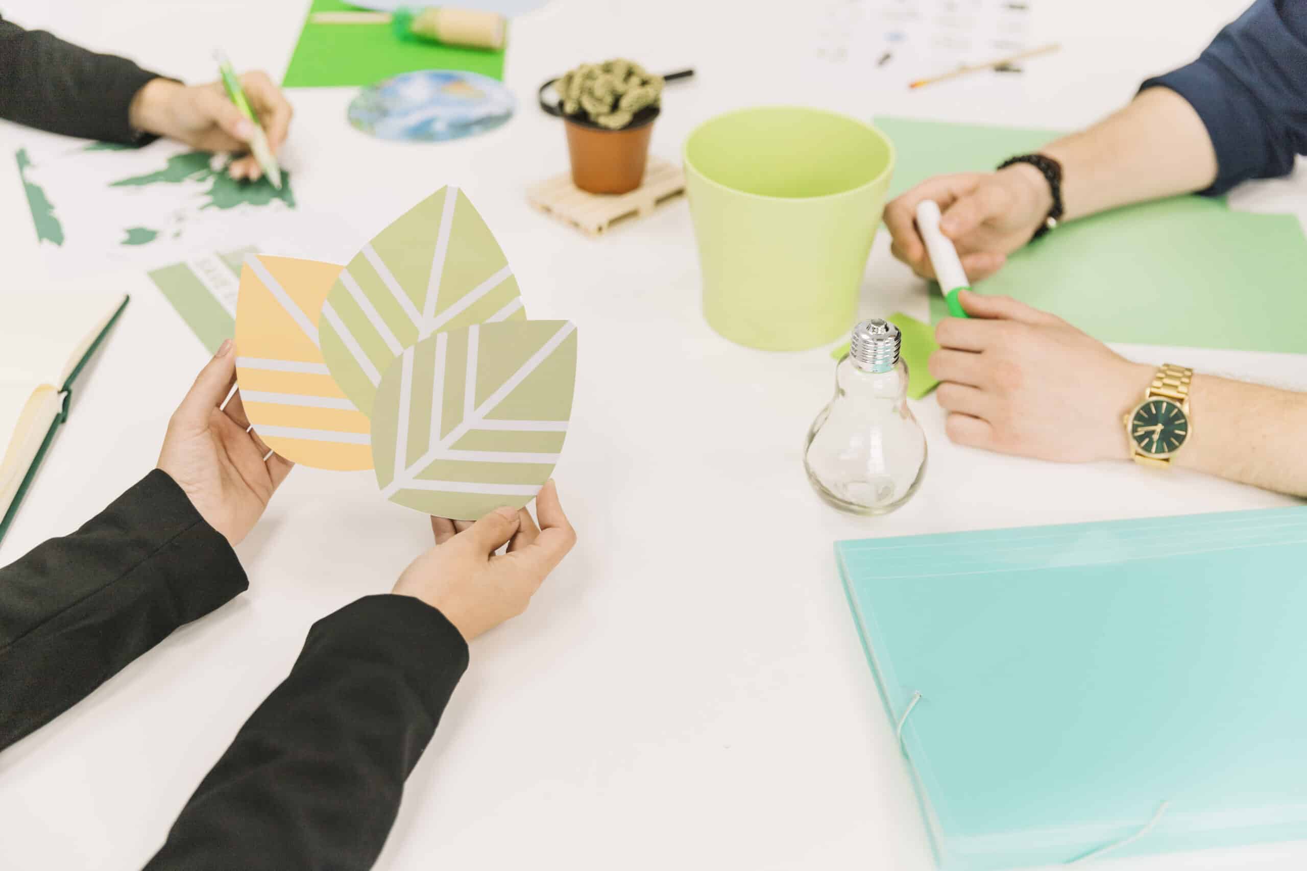 Pessoas reunidas em uma mesa de trabalho com elementos ecológicos, segurando ícones de folhas, lâmpada e materiais verdes, representando o design de produtos sustentáveis no ambiente digital.