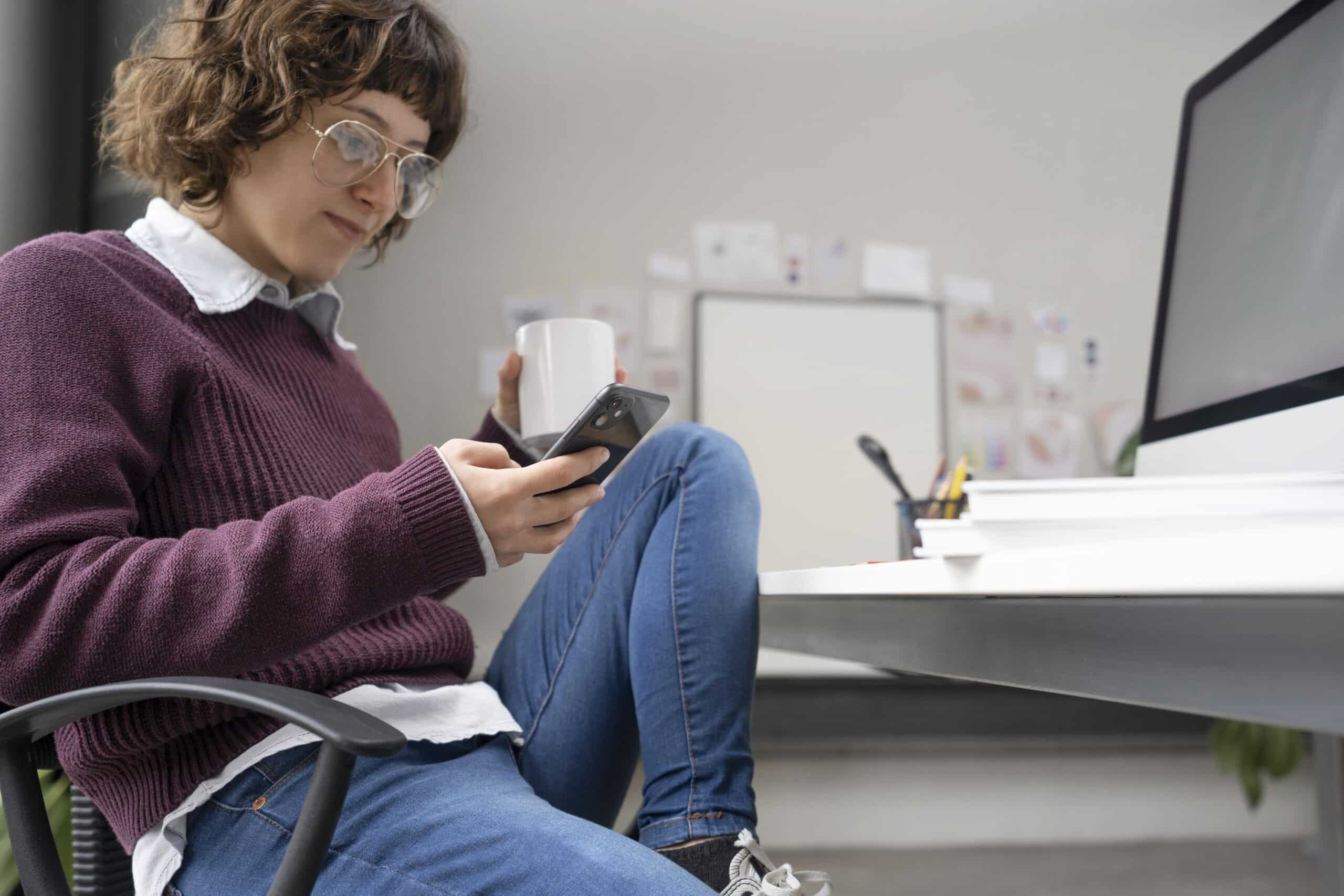 Jovem designer consultando informações no smartphone enquanto trabalha em seu escritório ao lado de um computador
