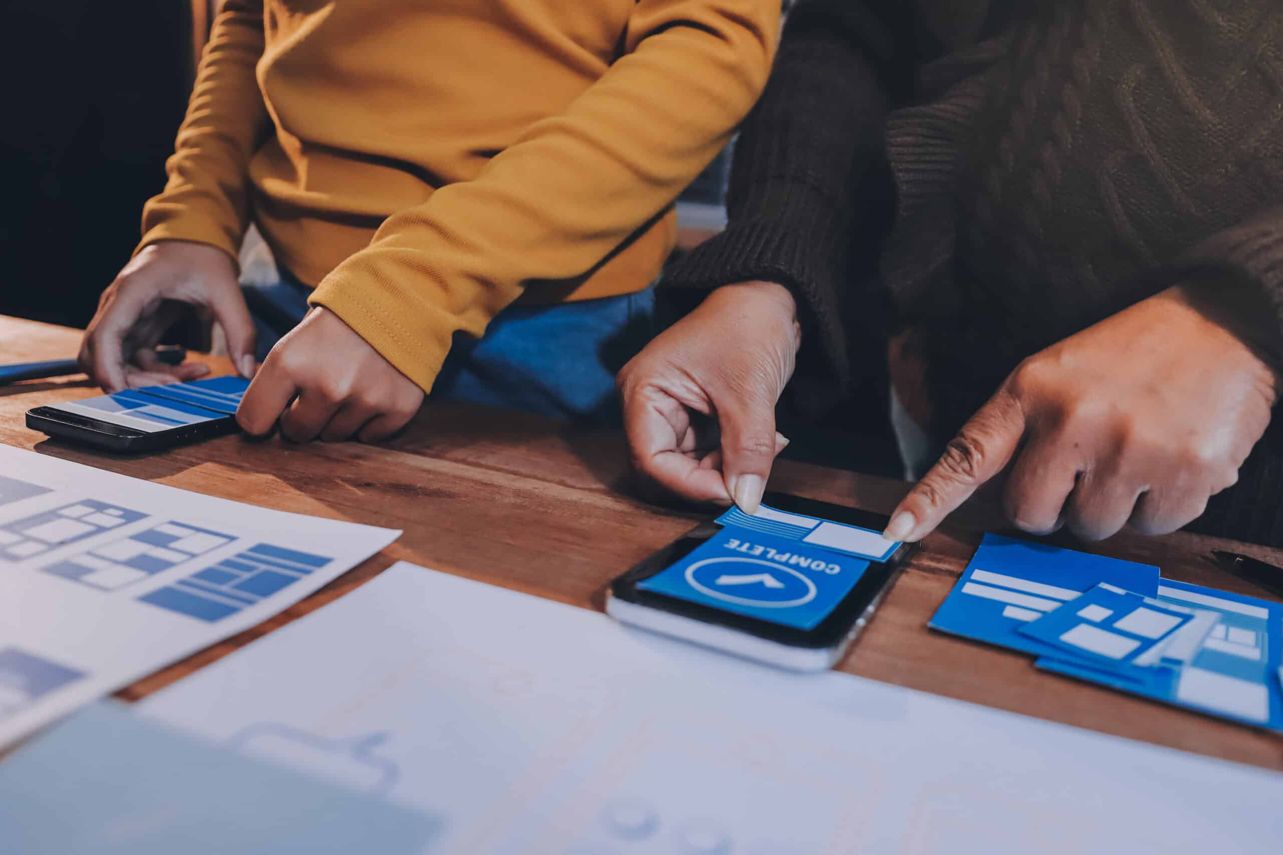 Duas pessoas trabalhando em protótipos de aplicativos móveis com elementos de design de interface sobre a mesa, representando o processo de UX e desenvolvimento digital.