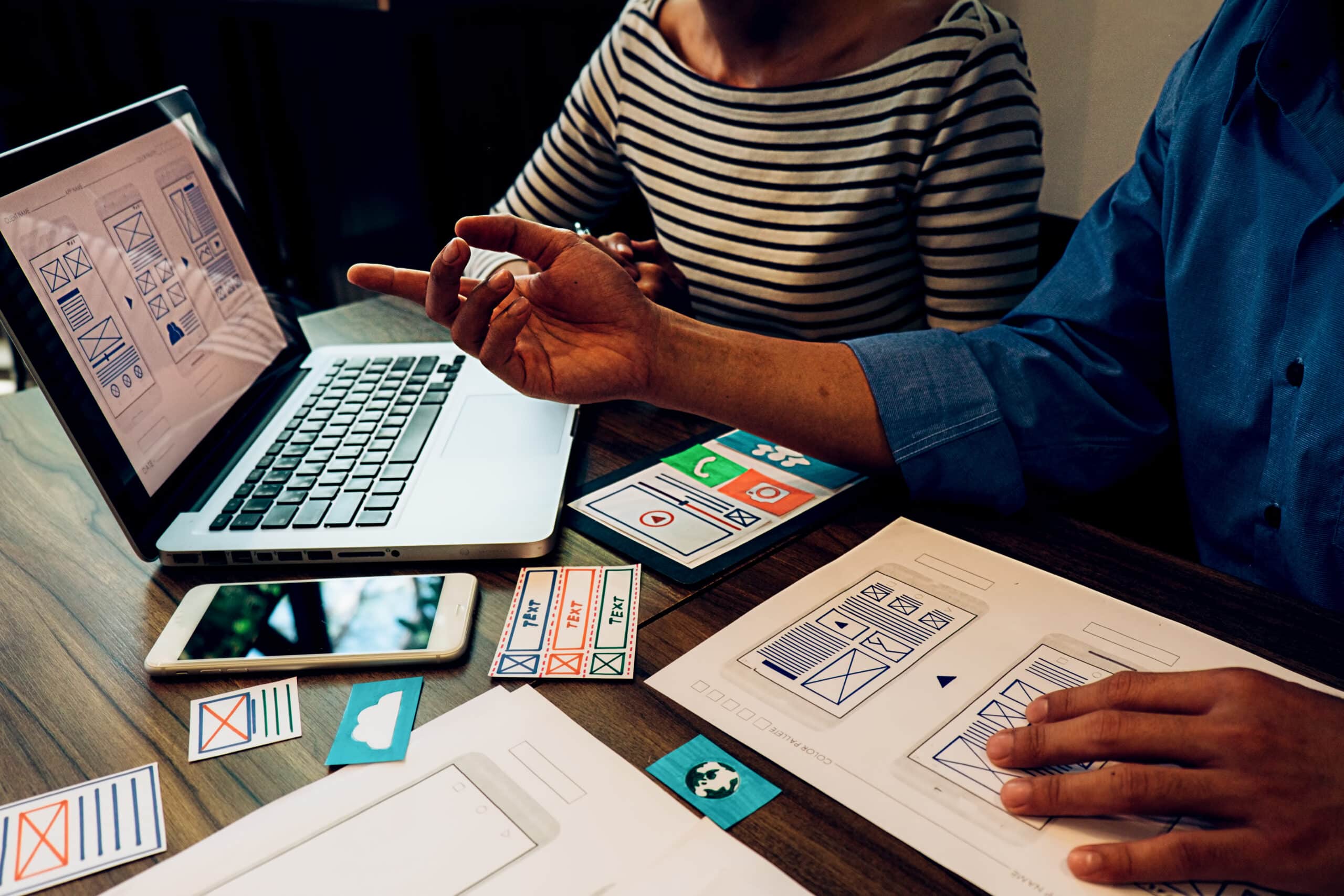 Duas pessoas colaborando em um projeto de design de conteúdo, analisando wireframes e protótipos em um laptop e em papéis sobre a mesa.